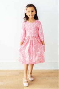 Girl wearing a pink 3/4 sleeve twirl dress with white hearts, side pockets, and ballet flats. Perfect for school or play.