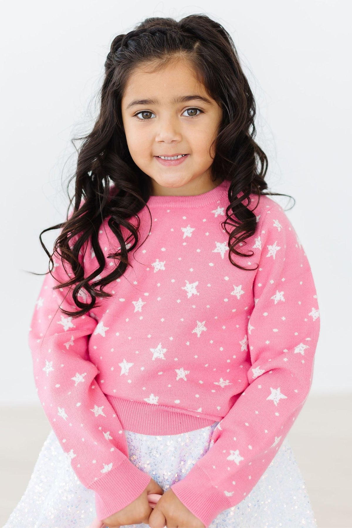 Child wearing pink Shining Star Sweater with white star pattern, layered over a sparkly silver sequin skirt for a cozy, festive look.