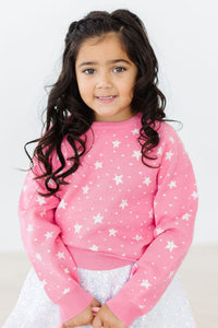 Child wearing pink Shining Star Sweater with white star pattern, layered over a sparkly silver sequin skirt for a cozy, festive look.