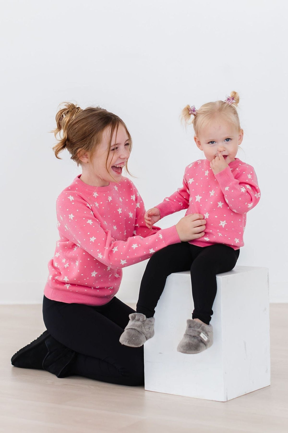 Two girls wear matching pink Shining Star Sweaters with white stars and black leggings for cozy chilly days.