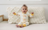 Baby wearing GOTS certified organic cotton zip romper with smiley face print and yellow cuffs, sitting on textured white pillows.