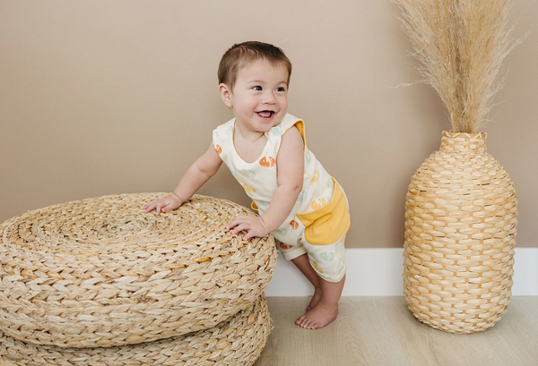 Happy baby wearing a GOTS certified organic cotton sleeveless romper with smiley bolts and yellow pockets, leaning on a woven stool.