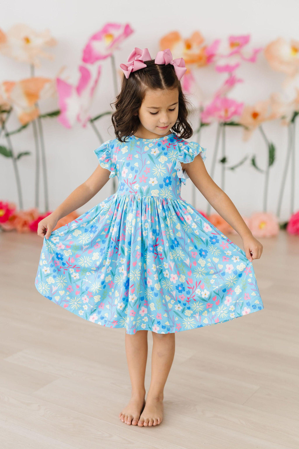 A child wearing the Springtime Splendor Flutter Sleeve Twirl Dress, showcasing a playful dance pose. The dress features a bright blue fabric with floral prints, complemented by charming flutter sleeves. Perfect for springtime, the dress embodies fun and elegance, ideal for joyful outings.