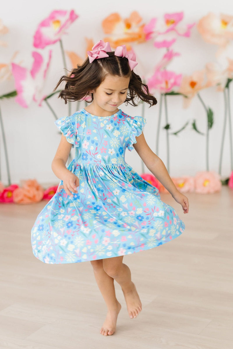 A joyful girl twirling in the Springtime Splendor Flutter Sleeve Twirl Dress, showcasing vibrant flowers against a blue background.
