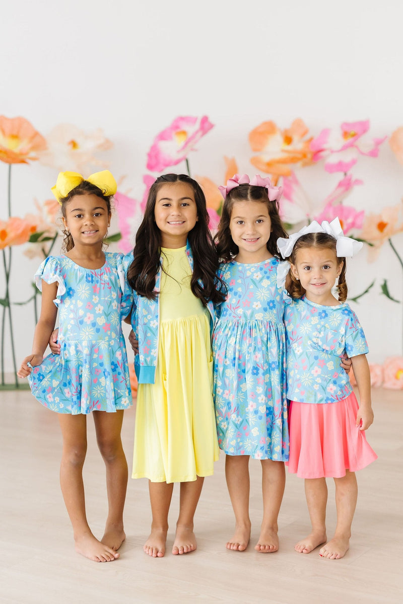 Four children in colorful outfits, including the Springtime Splendor Satin Jacket, posing together against a backdrop of vibrant flowers, showcasing spring fashion.