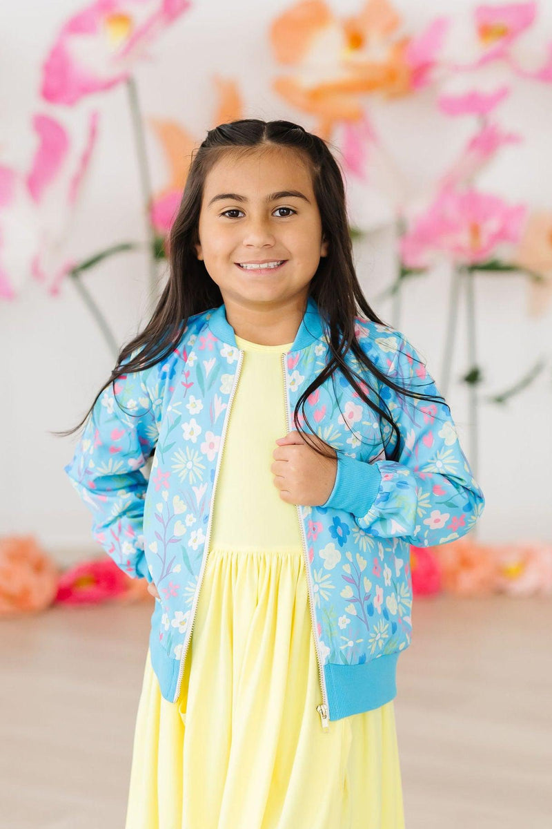 A happy girl wearing the vibrant Springtime Splendor Satin Jacket from Mila & Rose with a yellow dress, standing confidently against a backdrop of colorful flowers.