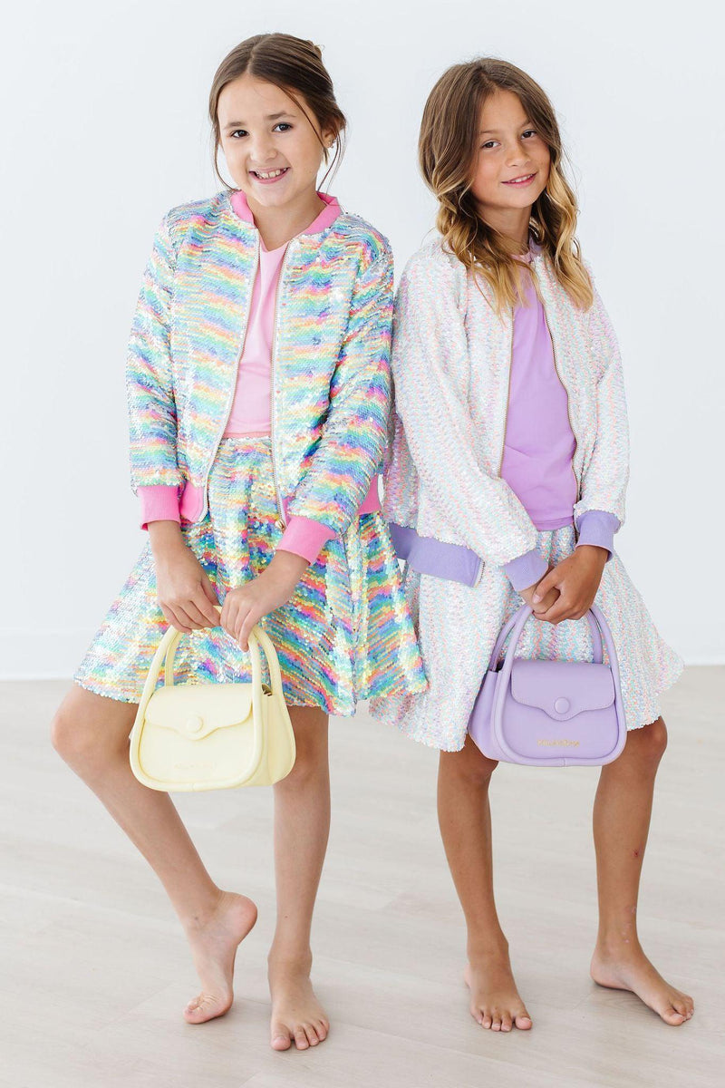 Two girls wearing the Sprinkles Flip Sequin Jacket by Mila & Rose®, each holding stylish handbags. Their outfits consist of colorful sequined jackets and skirts, radiating fun and youthful charm in a playful setting.