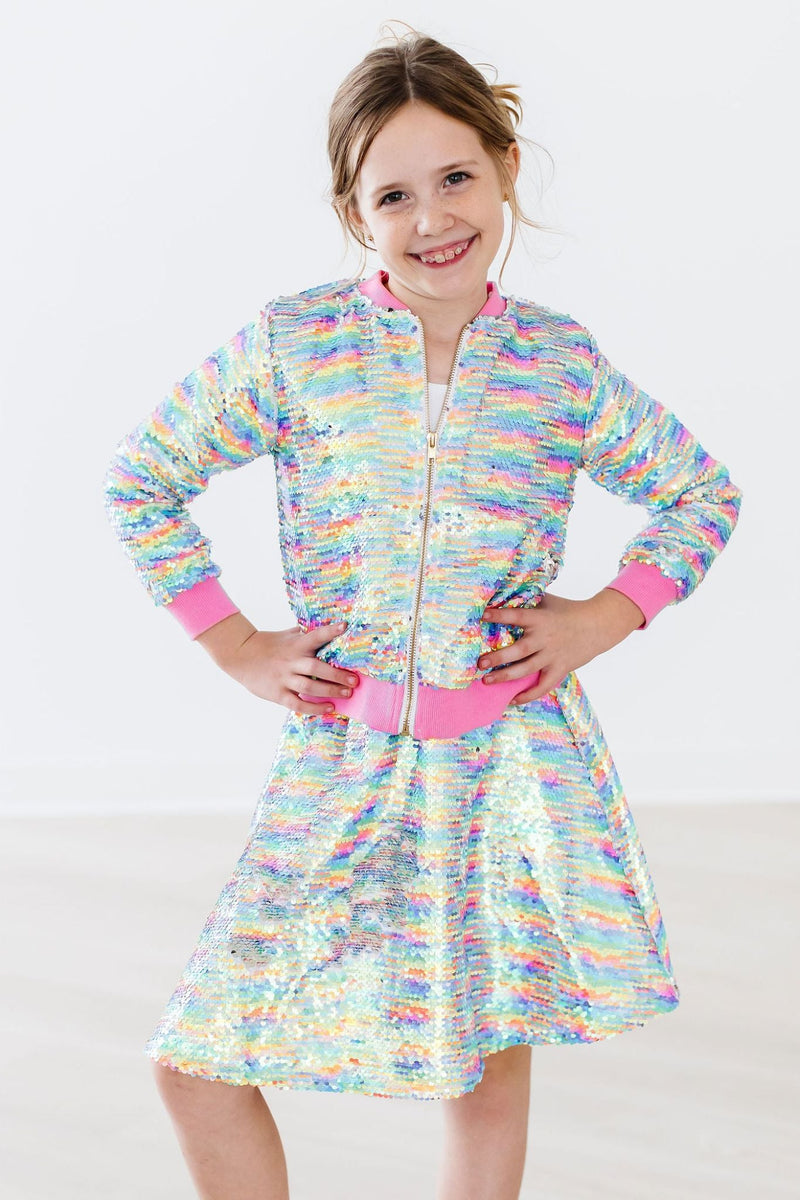 A smiling girl showcasing the Sprinkles Flip Sequin Jacket by Mila & Rose®, paired with a beautiful sequin skirt. The vibrant jacket features colorful sequins that change with a swipe, adding fun and glamour to her outfit.