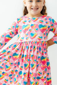 Smiling girl in Sugar & Spice pink dress with rainbow heart print, 3/4 sleeves, pockets, and a full twirl skirt.