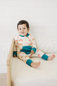 Toddler smiling in cream organic cotton playsuit with sunrise print and teal pocket, sitting on a bed.