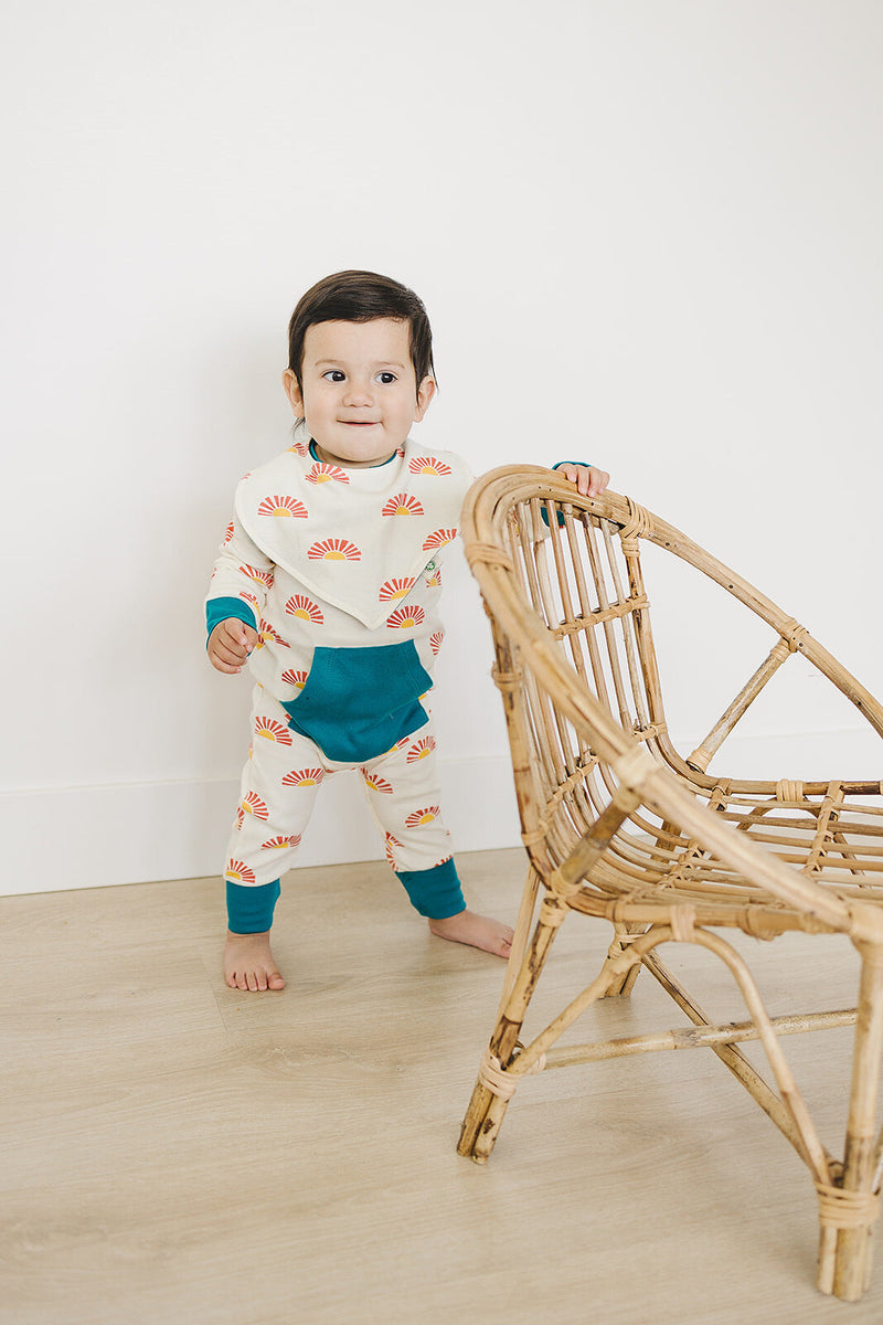 Baby standing by rattan chair wearing GOTS organic cotton playsuit with Sunrise print and teal pocket.