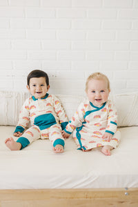 Two smiling babies sit on a bed wearing GOTS certified organic cotton playsuits with a sunrise print and teal trim.