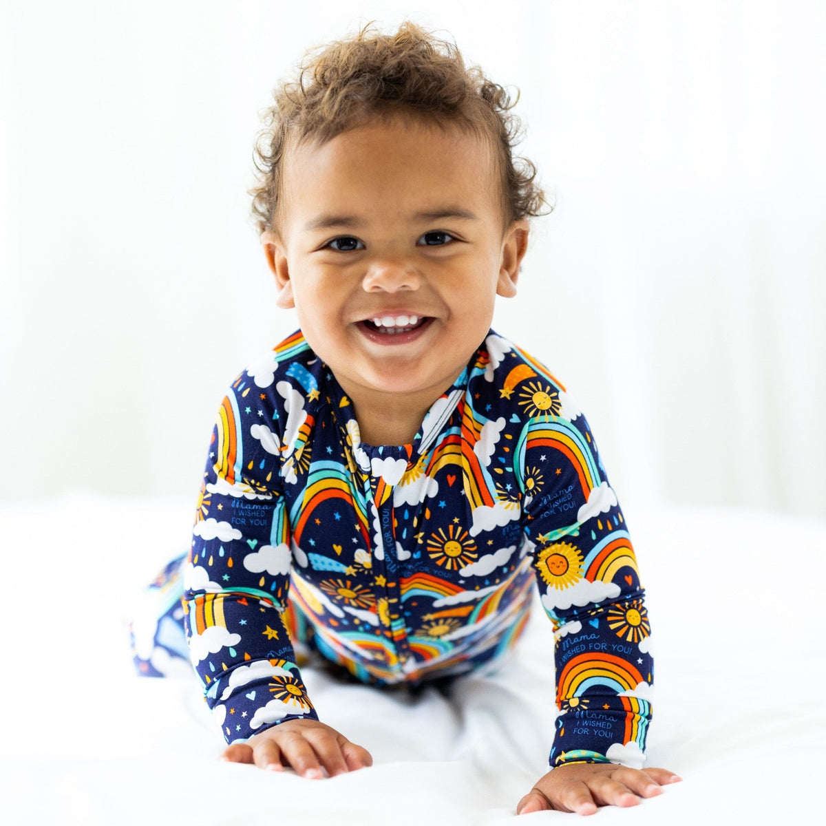 Smiling toddler wearing Here Comes the Sunshine blue Second-Skin Convertible Romper with sun, clouds, and rainbows.
