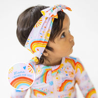 The SoftKnot Bow Here Comes the Sunshine Pink head-wrap bow on a child with pastel sun and rainbow print.