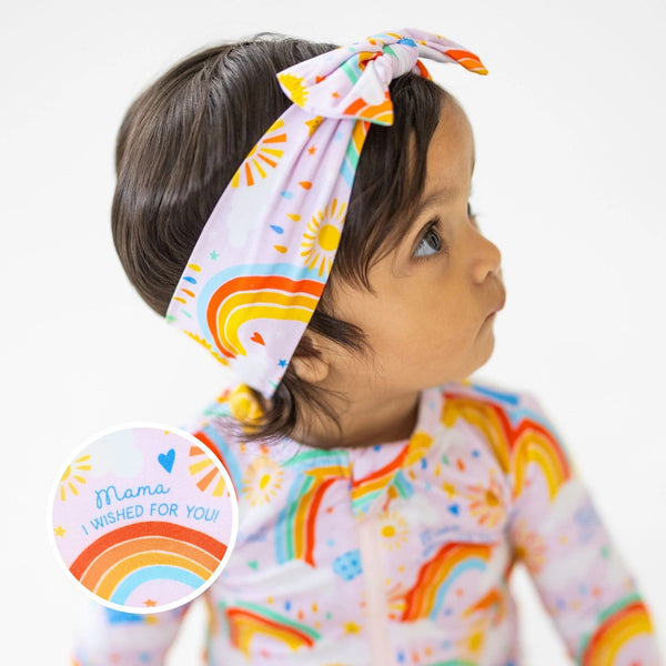 The SoftKnot Bow Here Comes the Sunshine Pink head-wrap bow on a child with pastel sun and rainbow print.