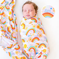 The Forever Swaddle Blanket - Here Comes the Sunshine (Pink) featuring a baby peacefully swaddled in a colorful rainbow-patterned blanket with suns and clouds, promoting comfort and joy.