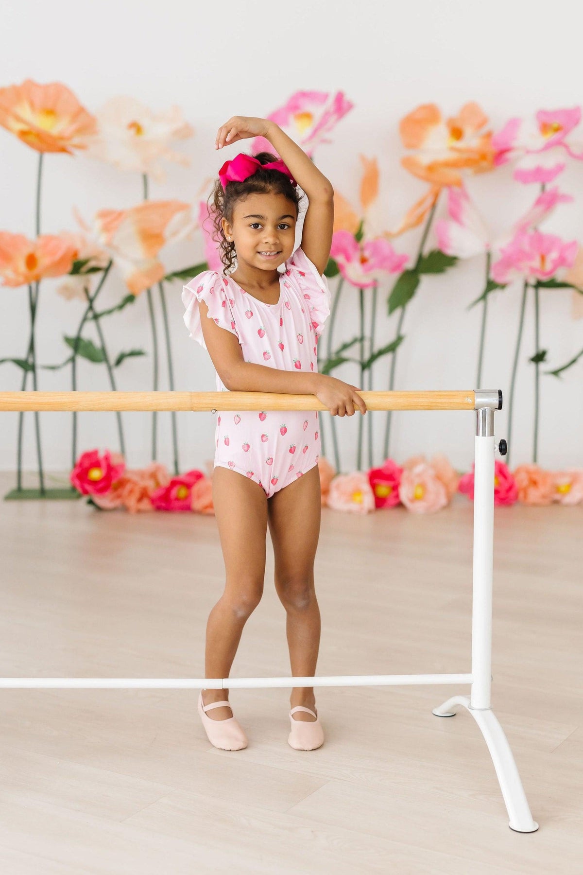 Tiny ballerina in Sweet Strawberries flutter-sleeve leotard at barre, pink bow, blooming backdrop.