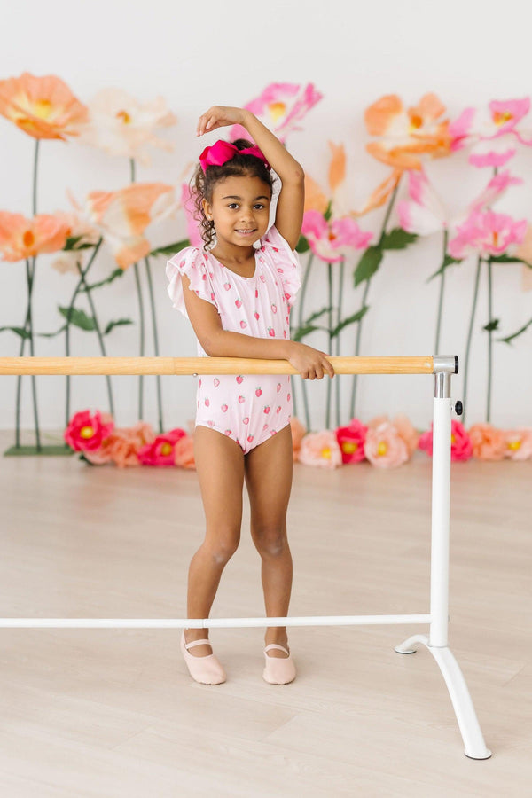 Tiny ballerina in Sweet Strawberries flutter-sleeve leotard at barre, pink bow, blooming backdrop.