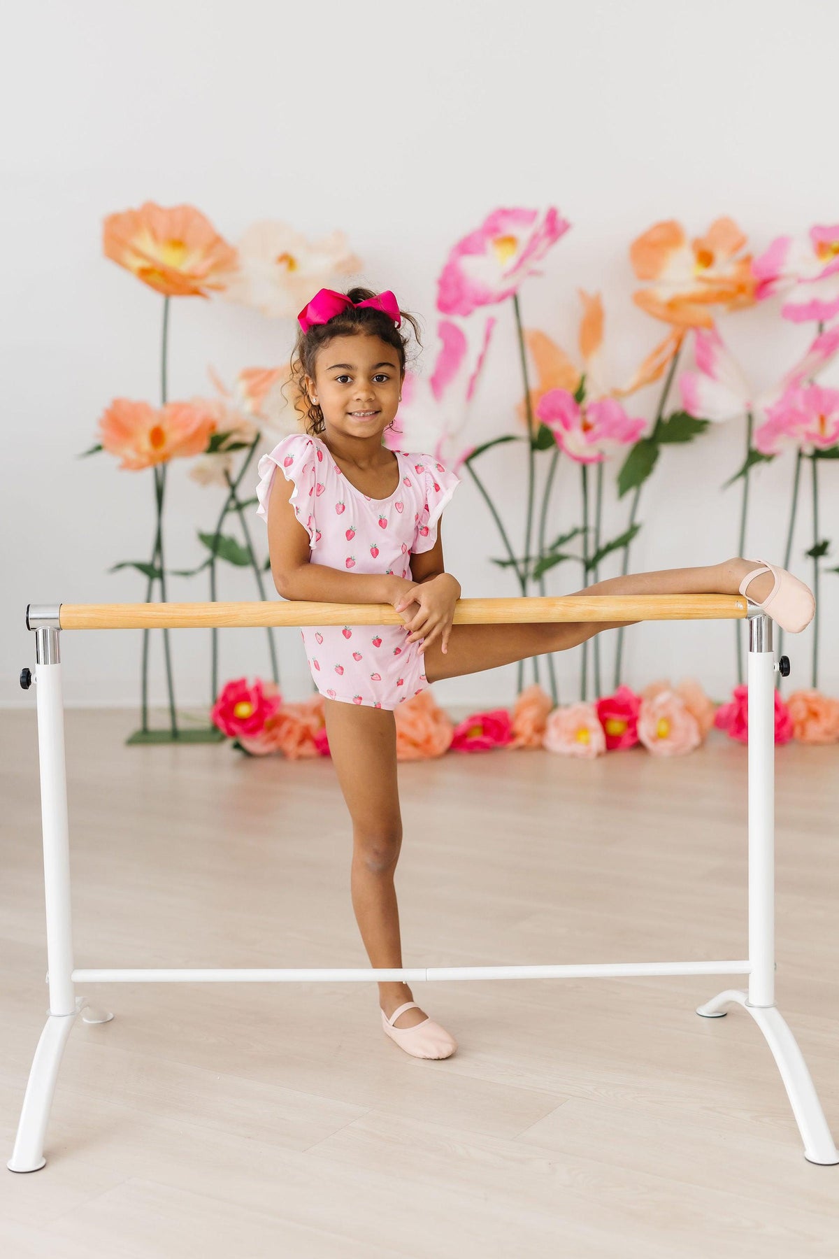 Young girl in Sweet Strawberries short sleeve flutter-sleeve pink leotard at a ballet barre among paper flowers.