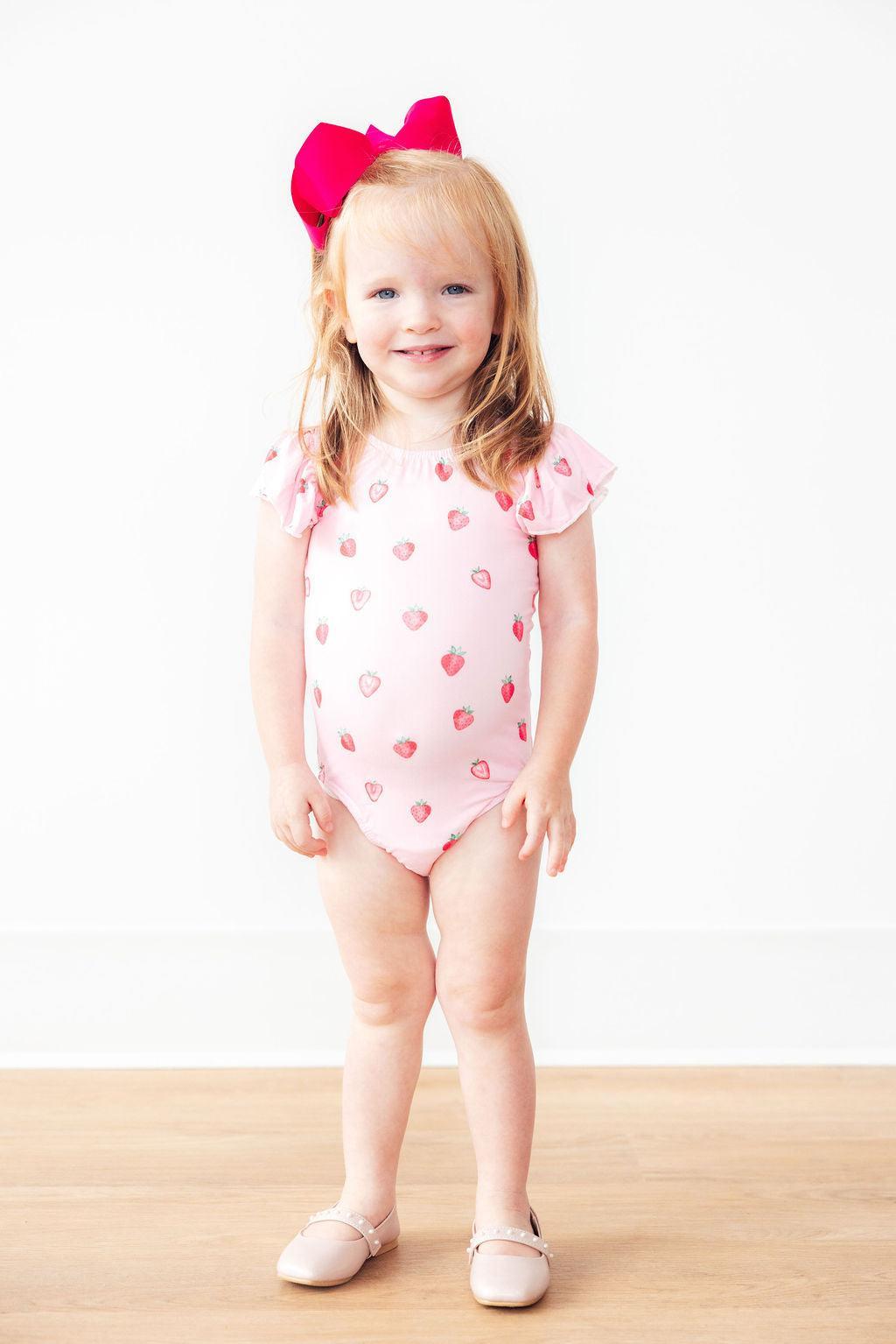 Sweet Strawberries short sleeve flutter sleeve leotard on a smiling toddler girl with a pink bow.