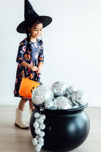 Girl in Halloween pumpkin print twirl dress and witch hat holding orange bucket beside cauldron filled with disco balls.