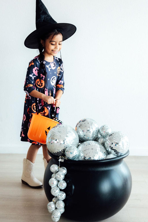Girl in Halloween pumpkin print twirl dress and witch hat holding orange bucket beside cauldron filled with disco balls.