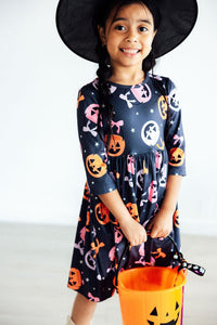Smiling girl in navy 3/4 sleeve twirl dress with pink and orange pumpkin print, witch hat, and orange Halloween candy bucket.