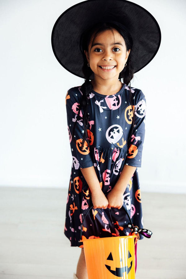 Smiling girl in a Halloween pumpkin print twirl dress and witch hat holding an orange candy bucket.