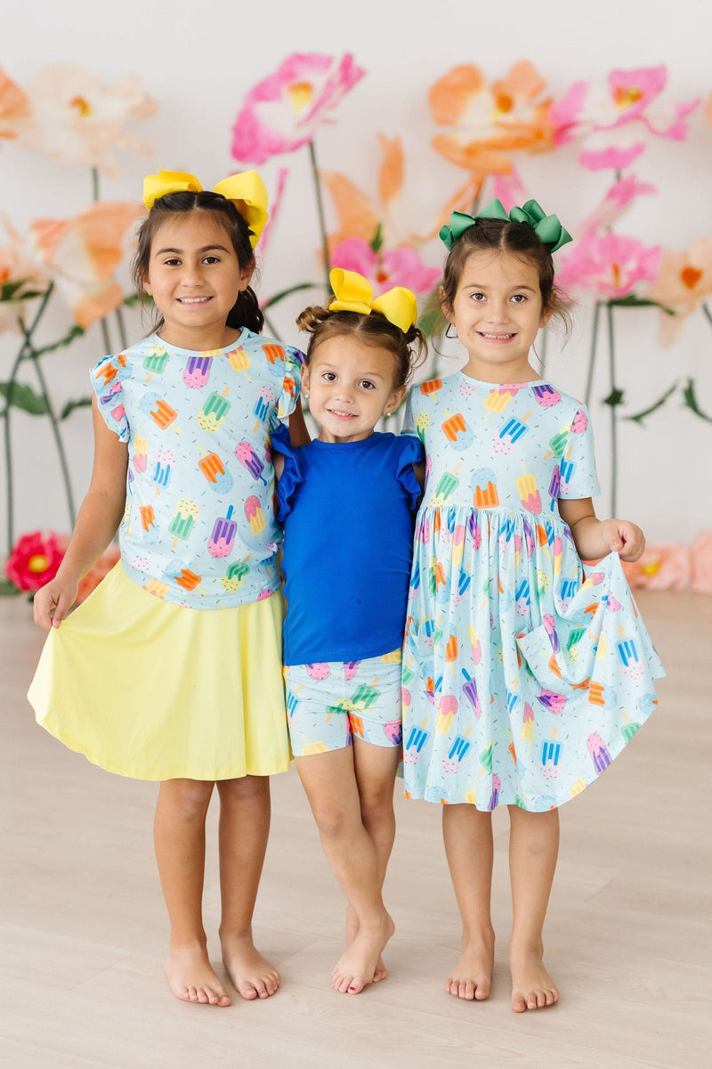 Three young girls showcase their Sweet Treat S/S Pocket Twirl Dresses in vibrant colors. The girls have fun hairstyles and are posing joyfully in a playful setting, illustrating the dress's comfort and style.