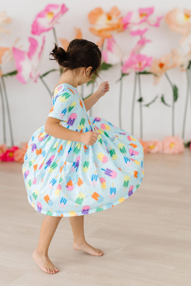 A child twirling in the Sweet Treat S/S Pocket Twirl Dress with a vibrant popsicle design, highlighting the dress's playful movement.