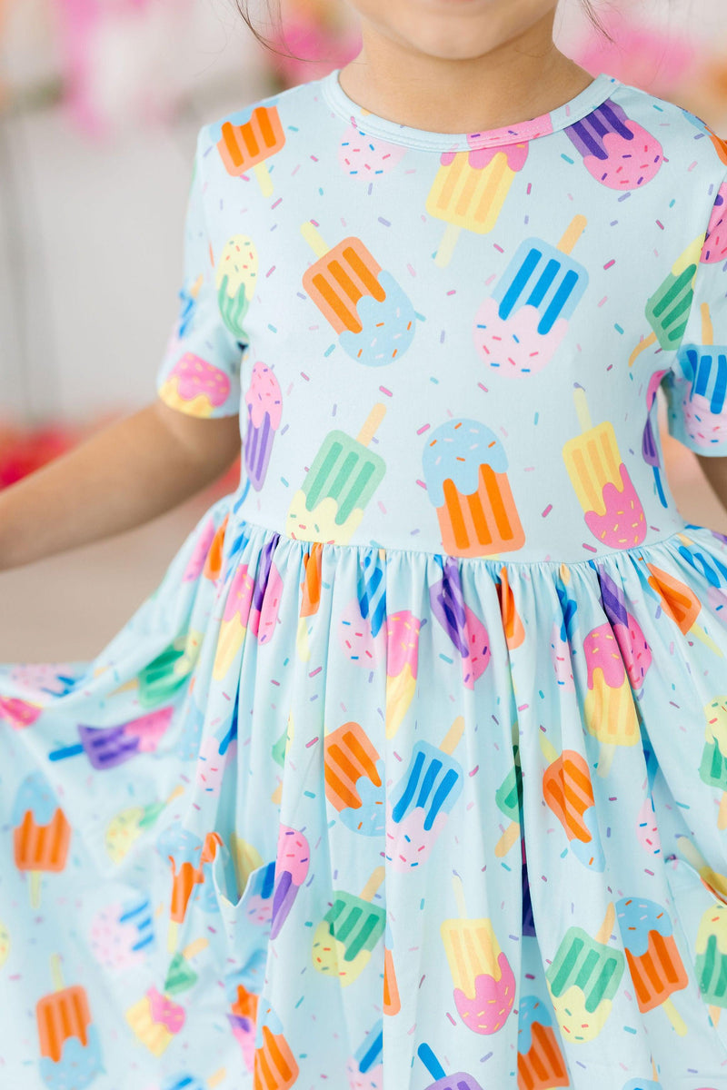 Close-up of the Sweet Treat S/S Pocket Twirl Dress displaying the fun popsicle pattern, held by a child showcasing its comfort and style.