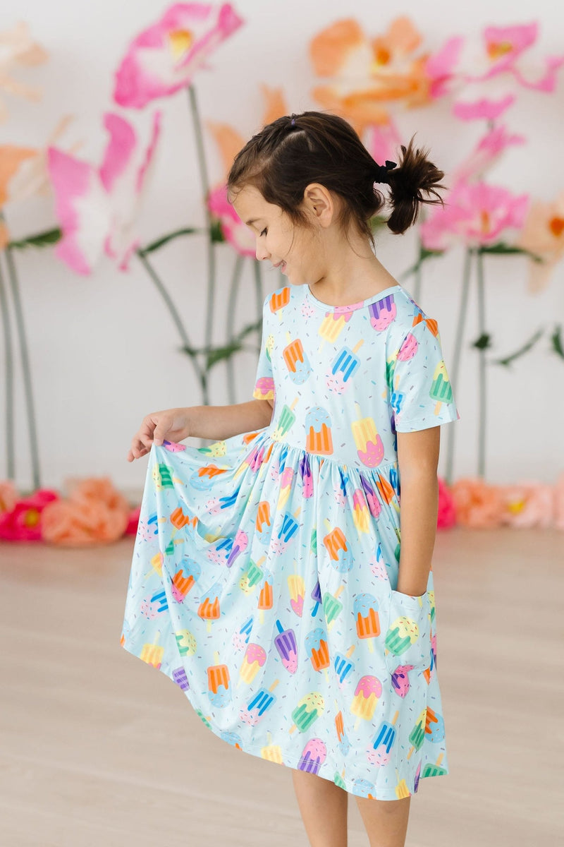A young girl admires the twirl of her Sweet Treat S/S Pocket Twirl Dress, featuring a playful ice cream pattern. The dress has a scoop back and convenient pockets, perfect for kids who love to play.