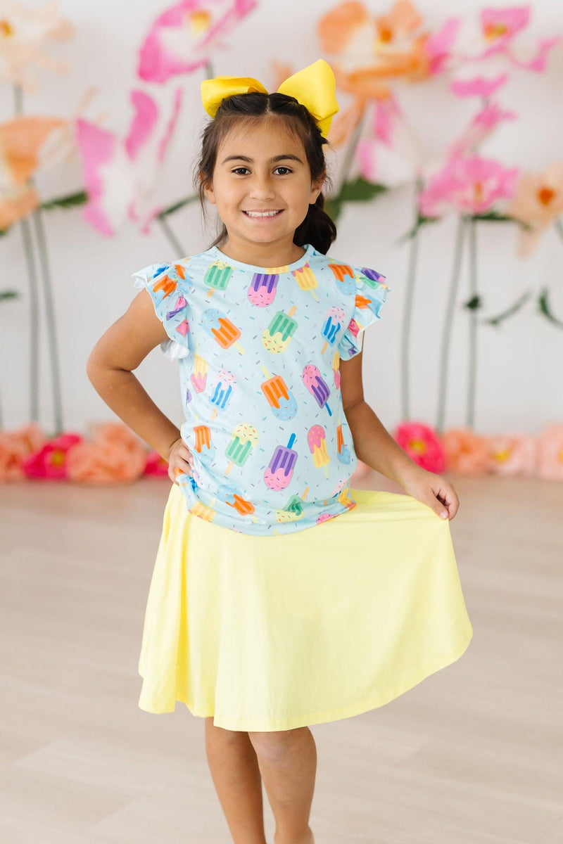 Girl joyfully showcasing the Adorable 'Sweet Treat' Ruffle Sleeve Tee paired with a yellow skirt, embodying playful style and comfort.