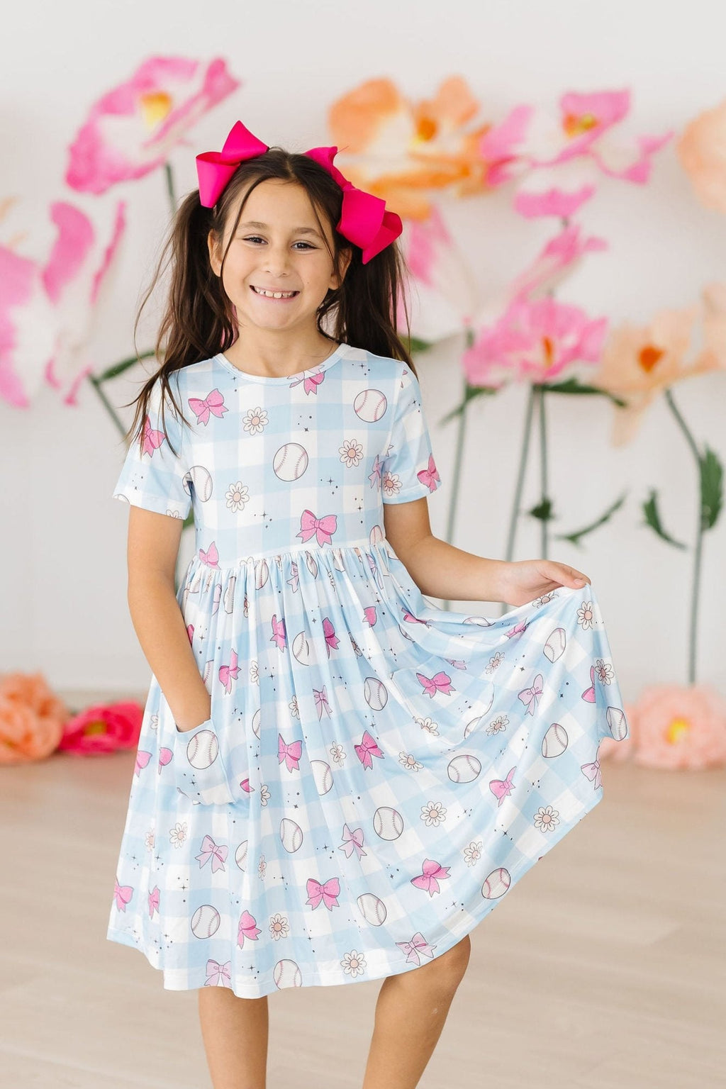 Take Me Out to the Bow Game S/S Pocket Twirl Dress shown on a smiling girl, featuring a playful blue pattern with baseballs and bows, perfect for twirling and playtime.