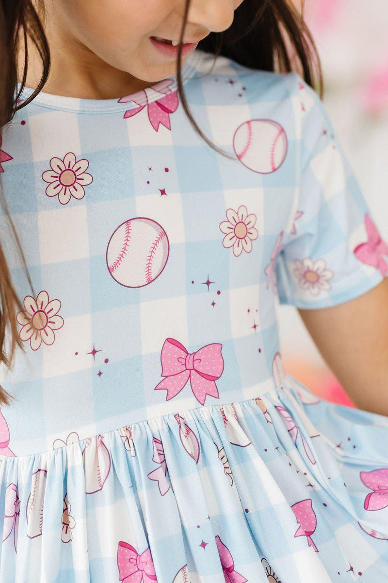 Close-up of the Take Me Out to the Bow Game S/S Pocket Twirl Dress, highlighting the cute blue gingham design with baseballs, cheerful bows, and floral accents.