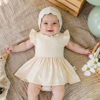 Baby lying on beige blanket wearing sand twirl bodysuit dress with cream eyelet bonnet and props