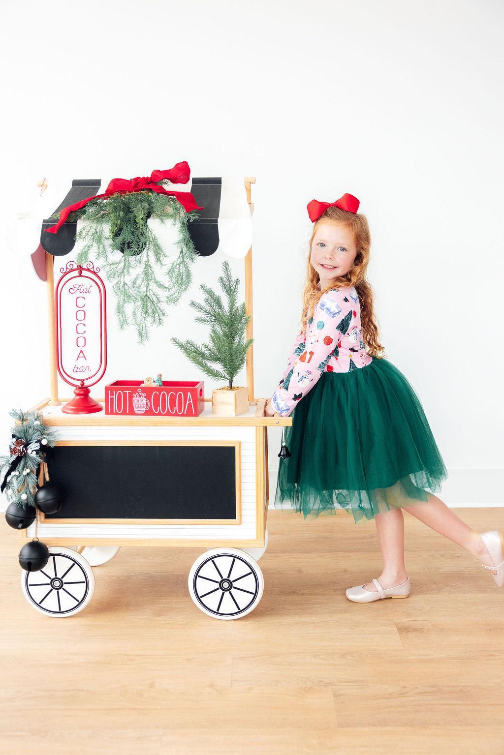 Girl in long sleeve tutu dress with red bow posing next to a festive hot cocoa cart decorated with evergreen branches.
