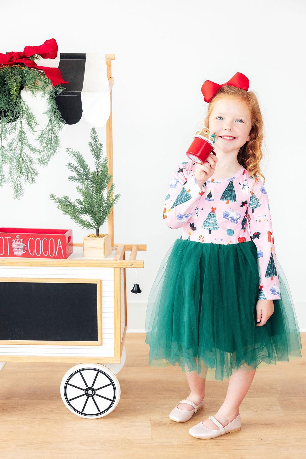 Girl wearing pink Christmas print long sleeve tutu dress with green skirt and red bow, holding a mug near a hot cocoa cart.