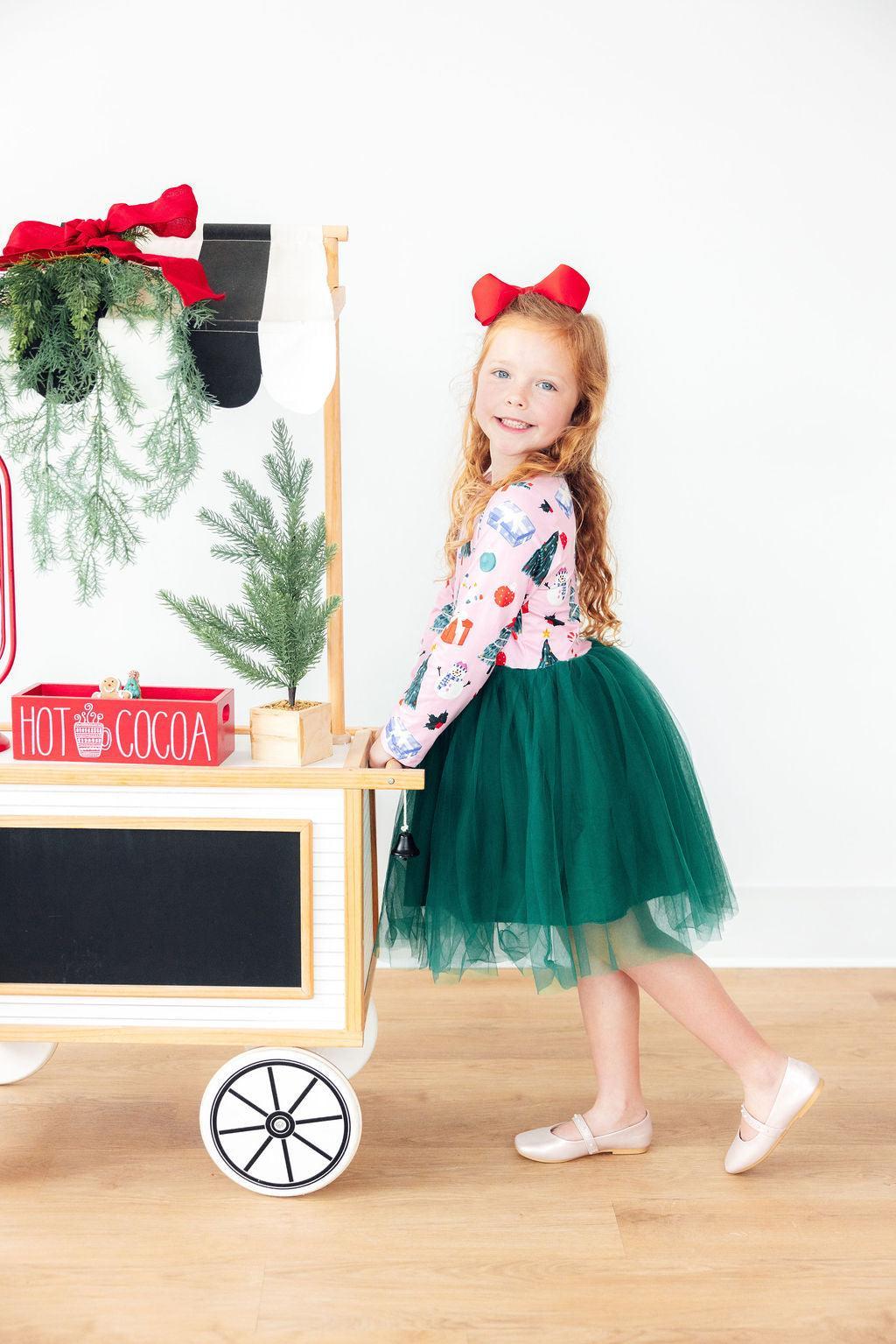 Little girl in festive long sleeve tutu dress with green tulle skirt and red bow, posing near holiday cart.