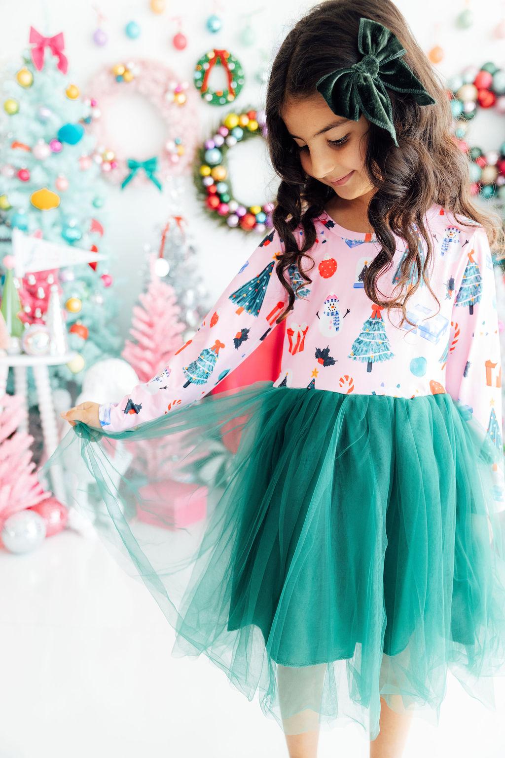 Girl wears pink long sleeve tutu dress with Christmas print and green skirt, holding the tulle against a festive holiday backdrop.