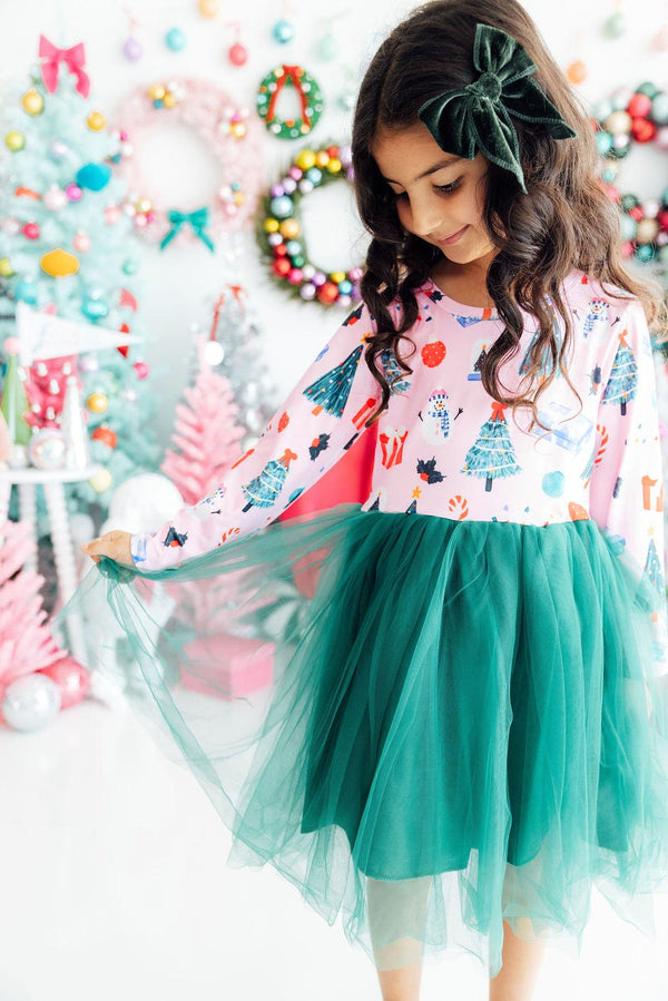 Girl wears pink long sleeve tutu dress with Christmas print and green skirt, holding the tulle against a festive holiday backdrop.
