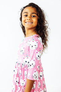 Smiling girl in pink Unicorn Kitties 3/4 sleeve pocket twirl dress with white cat/unicorn print.