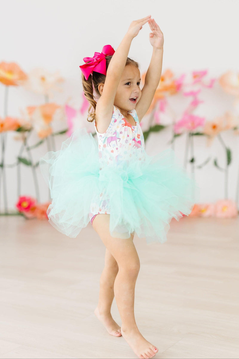 A joyful child wearing the Unicorn Wish Tank Tutu Leotard from Mila & Rose, showcasing a vibrant design with a fluffy mint tutu, dancing with excitement against a backdrop of colorful flowers.