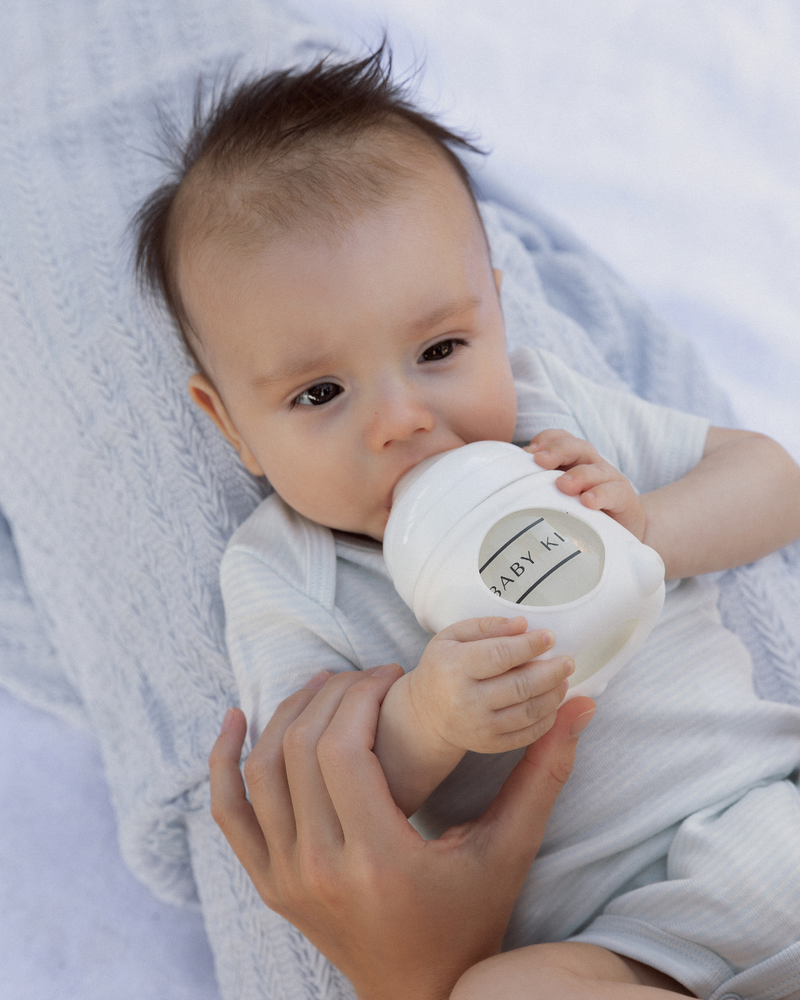 Baby holding a white bottle with 'BABY KY' branding, lying on a soft surface.