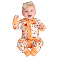 A smiling baby wearing a one-piece footed sleeper with a pumpkin and floral print, orange trim, and a matching beige bow headband, photographed against a plain white background.