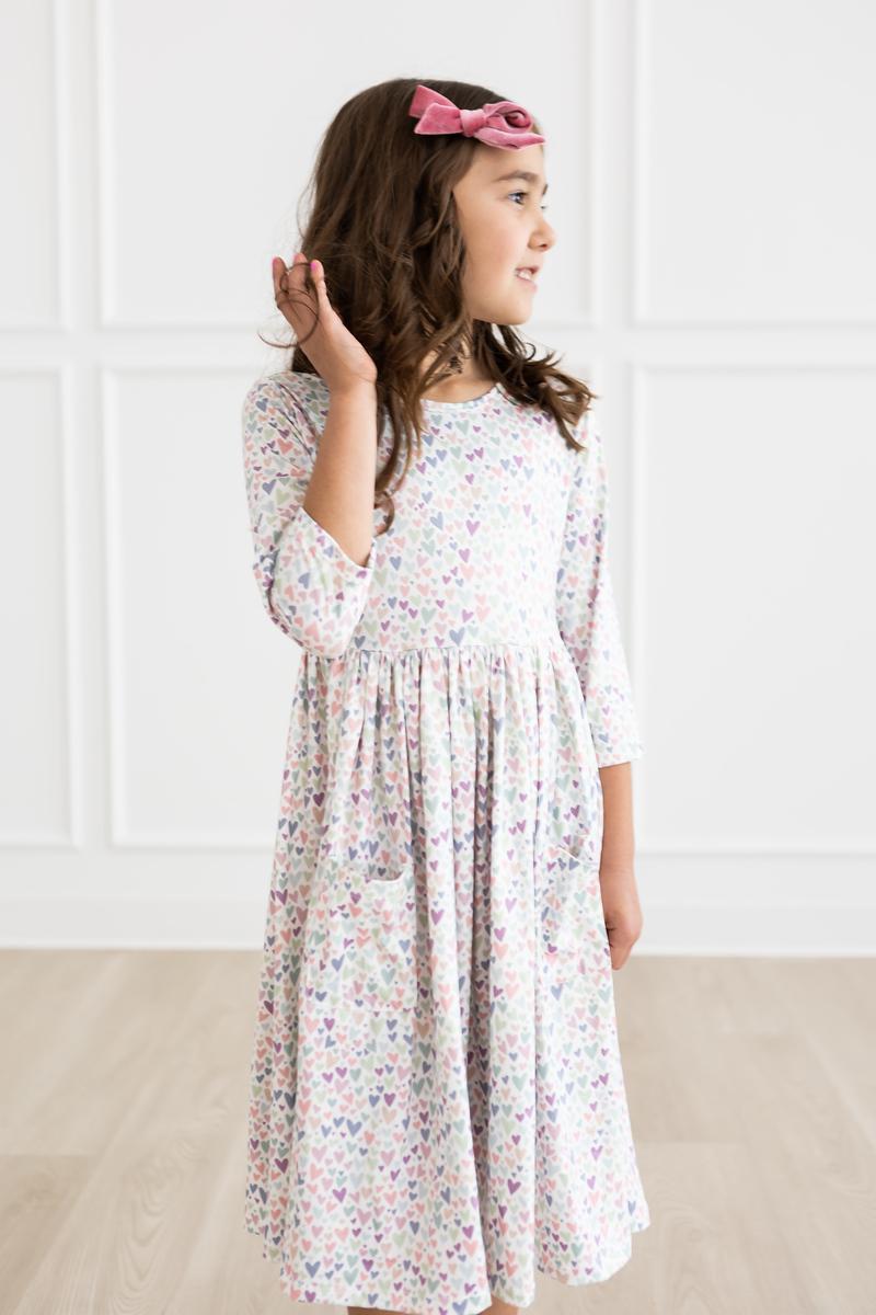 Little girl in pastel heart-print twirl dress with 3/4 sleeves, full skirt, and pink bow.