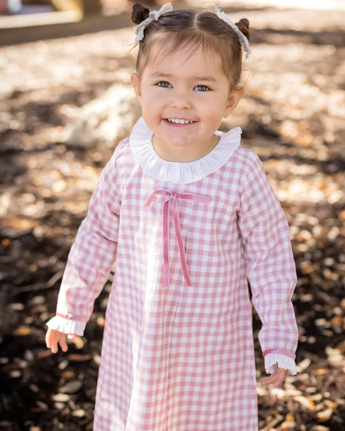 Smiling toddler wearing a pink gingham dress with a white ruffled collar, deep rose velvet bow, and matching ruffled cuffs.