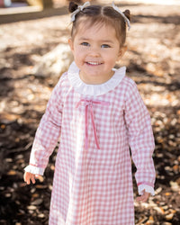 Smiling toddler wearing a pink gingham dress with a white ruffled collar, deep rose velvet bow, and matching ruffled cuffs.