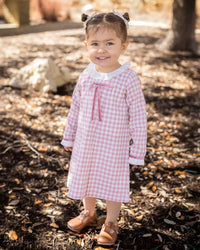 Girl wearing pink gingham A-line dress with white ruffled collar, darker rose velvet bow at chest, and ruffled cuffs.