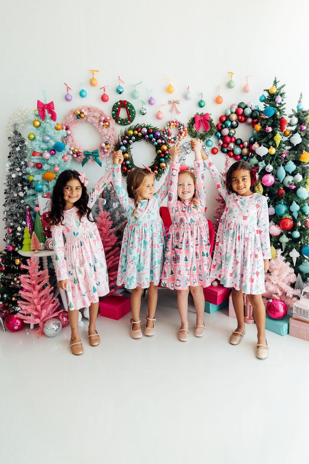 Four girls hold hands in festive long sleeve pocket twirl dresses amidst colorful Christmas trees and wreaths.